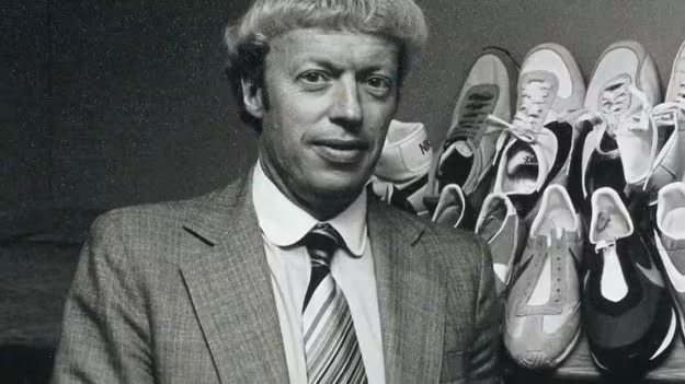 Phil Knight standing beside a display of Nike running shoes.